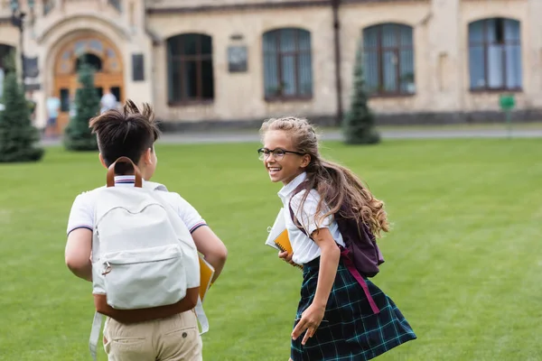 Positive schoolkid in eyeglasses running near asian friend with backpack on lawn — Stock Photo