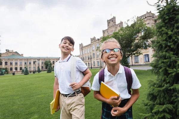 Fröhliche multiethnische Schulkinder mit Kopierbüchern blicken auf Rasen im Park in die Kamera — Stockfoto