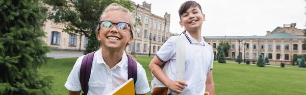 Positive schoolgirl in eyeglasses looking at camera near asian friend in park, banner — Stock Photo