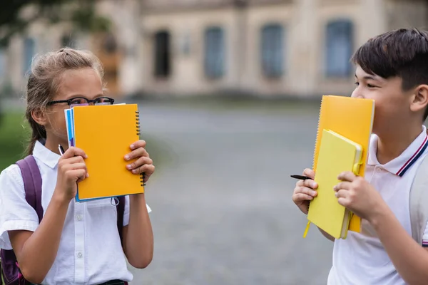 Мультиэтнические школьники держат блокноты рядом с лицами и смотрят друг на друга на улице — стоковое фото