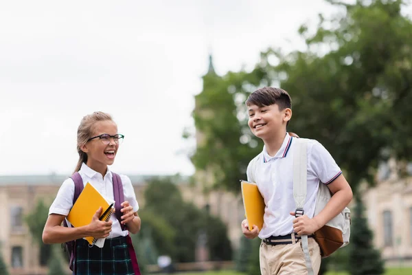 Positive interracial schoolchildren with notebooks holding backpacks outdoors — Stock Photo