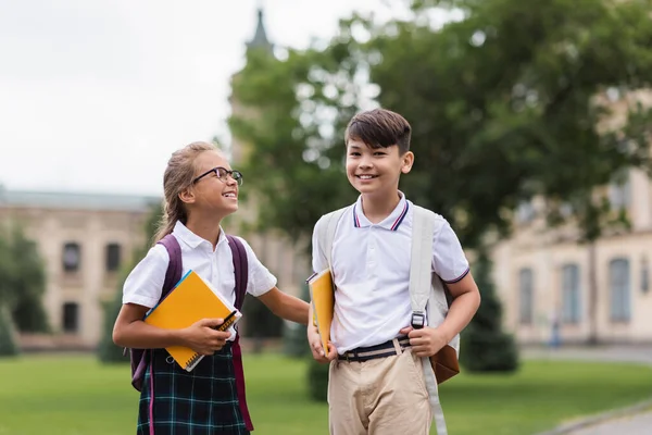 Colegiala en gafas celebración de cuadernos cerca asiático amigo al aire libre - foto de stock