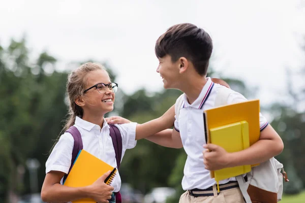 Fröhliche multiethnische Schulkinder mit Notizbüchern, die sich im Freien anschauen — Stockfoto