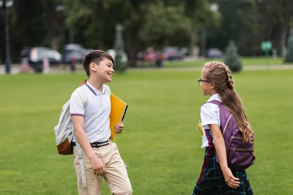 Lächelnde multiethnische Schulkinder mit Notizbüchern auf dem Rasen im Park — Stockfoto