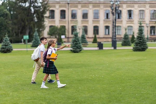 Vista laterale della studentessa che punta con il dito vicino all'amico asiatico con lo zaino mentre cammina nel parco — Foto stock