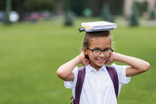 Smiling schoolgirl in eyeglasses holding notebooks on head outdoors — Stock Photo