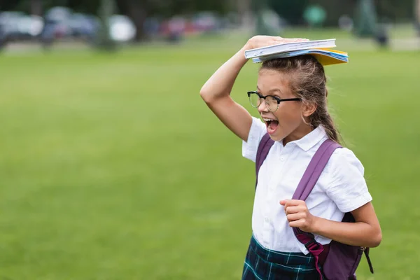 Aufgeregter Schüler mit Rucksack, der Kopierbücher auf dem Kopf im Freien hält — Stockfoto