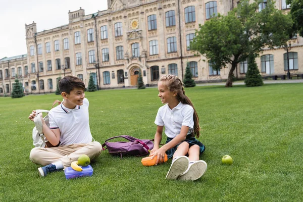 Sonrientes escolares multiétnicos sentados cerca de loncheras en el césped en el parque - foto de stock