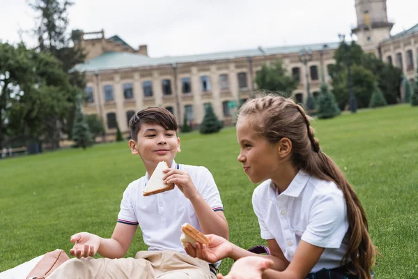 Усміхнений азіатський школяр тримає бутерброд біля друга на газоні в парку — стокове фото