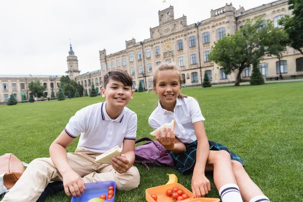 Lächelnde multiethnische Schulkinder halten Sandwiches in der Nähe von Lunchboxen auf dem Rasen im Freien — Stockfoto