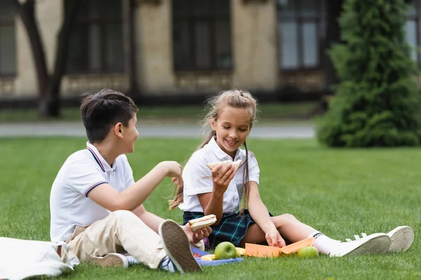 Positivo studentessa holding sandwich vicino asiatico compagno di classe su prato in parco — Foto stock