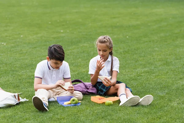Écoliers interraciaux tenant des sandwichs près des sacs à dos et des boîtes à lunch sur l'herbe — Photo de stock