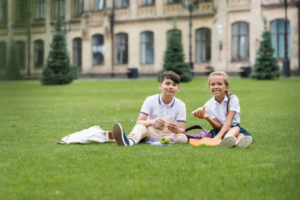 Cheerful interracial classmates holding sandwiches near lunchboxes on grass in park — Stock Photo
