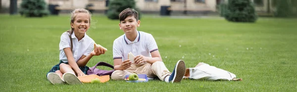 Positive gemischtrassige Kinder mit Sandwiches in der Nähe von Rucksäcken auf Gras im Park, Banner — Stockfoto