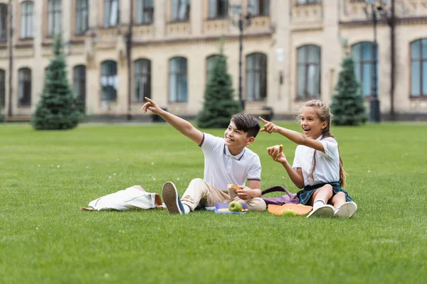 Sonrientes escolares interracial sosteniendo sándwiches y señalando con los dedos en el césped en el parque - foto de stock