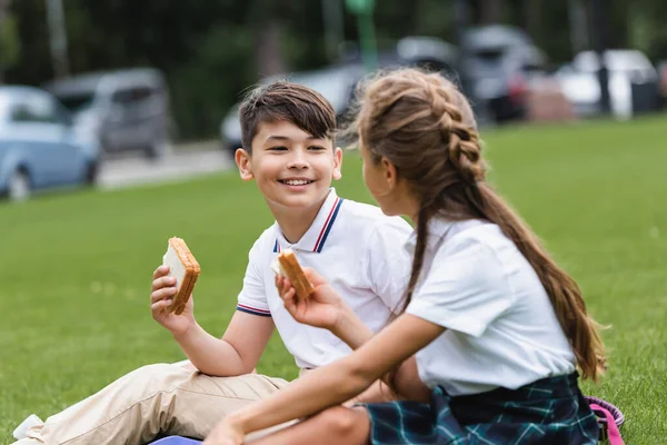 Smiling asian schoolboy holding sandwich near blurred friend on lawn in park — Stock Photo