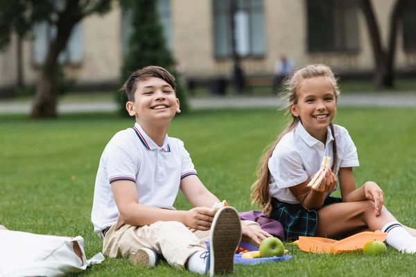 Positive multiethnische Schulkinder halten Sandwiches in der Nähe von Lunchboxen auf Gras im Freien — Stockfoto