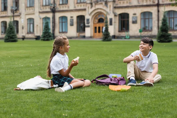 Multiethnic schoolkids holding sandwiches and talking on grass in park — Stock Photo