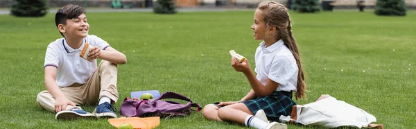 Школярка тримає сендвіч і розмовляє з азіатським другом на траві в парку, банер — стокове фото