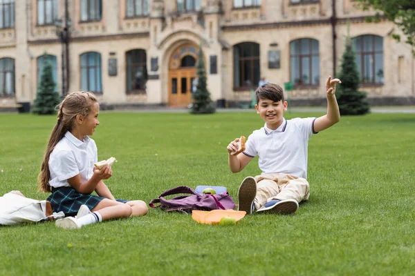 Asian schoolboy holding sandwich and pointing with finger near backpack and classmate in park — Stock Photo