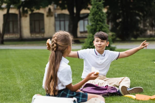 Asian schoolboy holding sandwich and talking to blurred friend on grass in park — Stock Photo
