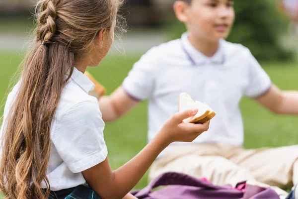 Preteen schoolgirl holding sandwich near blurred asian friend in park — Stock Photo