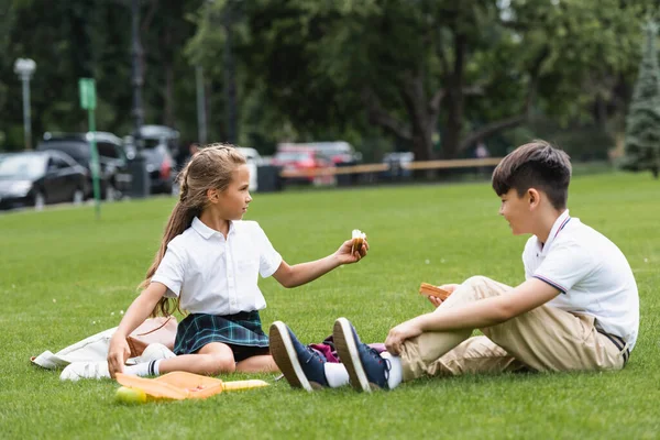 Vista laterale del bambino in età scolare che tiene il panino vicino al compagno di classe asiatico sull'erba nel parco — Foto stock