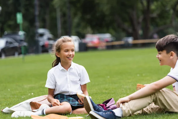 Счастливая школьница держит сэндвич рядом с размытым азиатским одноклассником, сидящим на траве в парке — стоковое фото