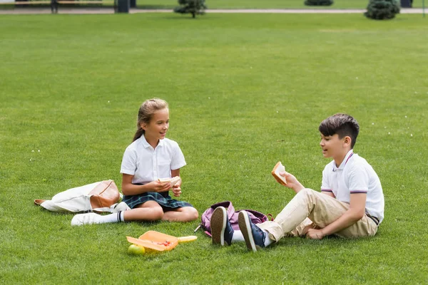 Multiethnic schoolchildren holding sandwiches near backpacks on grass in park — Stock Photo