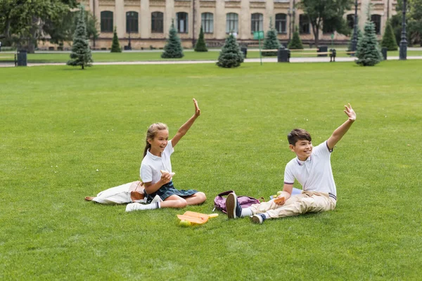 Fröhliche interrassische Schulkinder halten Sandwiches in der Hand und winken auf dem Rasen im Park — Stockfoto