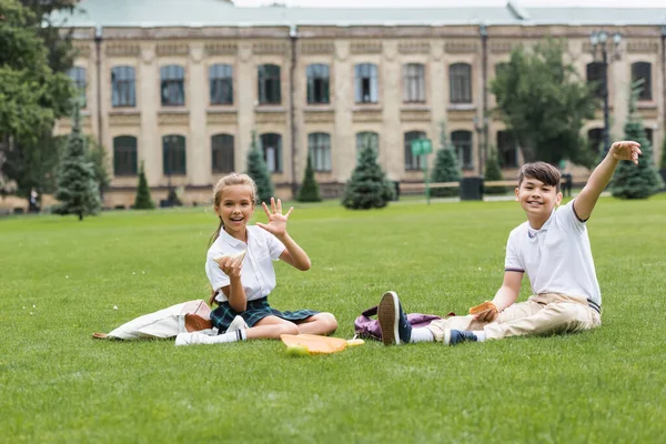 Positive schoolgirl holding sandwich and waving hand at camera near asian friend on lawn in park — Stock Photo