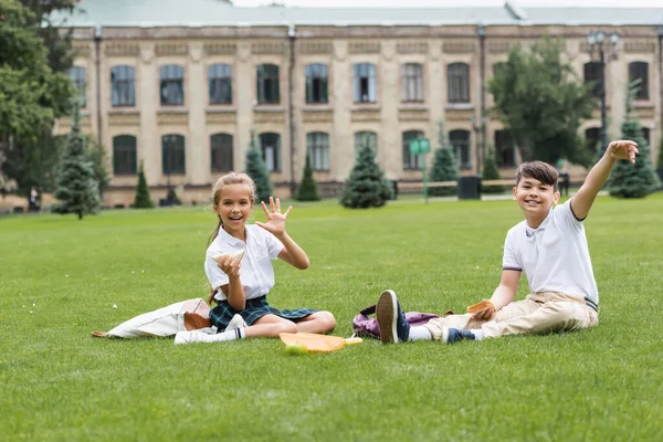 Positiva colegiala sosteniendo sándwich y agitando la mano en la cámara cerca de asiático amigo en el césped en parque - foto de stock