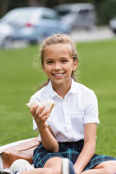 Smiling preteen schoolgirl holding sandwich on lawn in park — Stock Photo