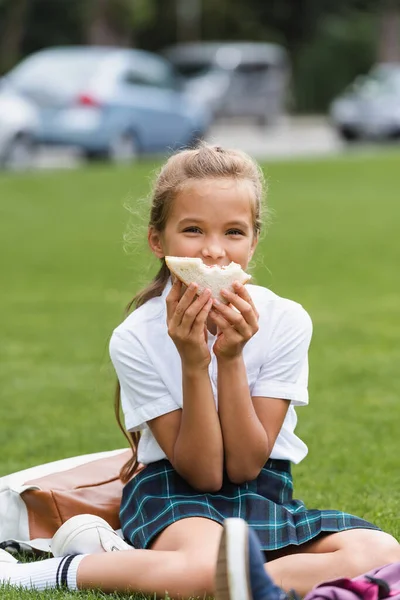 Школярка тримає сендвіч біля обличчя на траві в парку — стокове фото