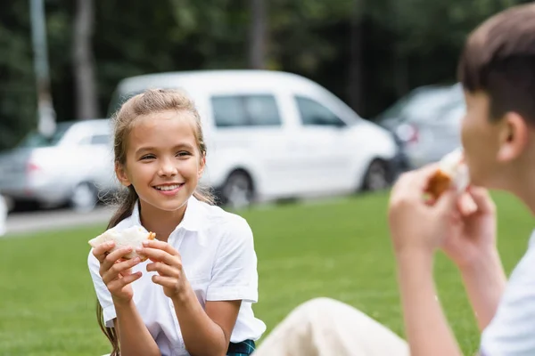 Smiling schoolgirl holding sandwich near blurred classmate in park — Stock Photo