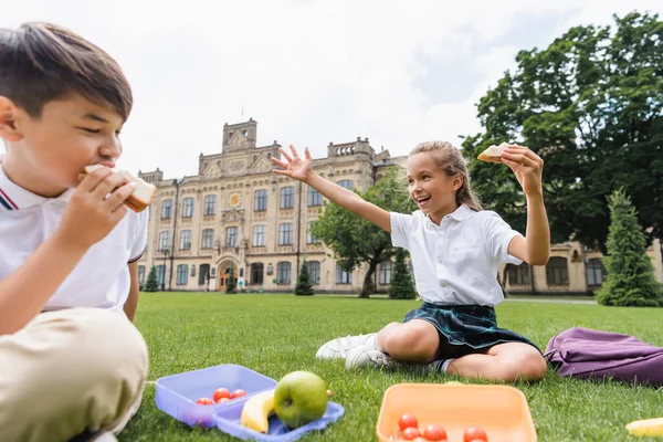 Studentessa holding sandwich e agitando mano vicino pranzo scatole e offuscata asiatico amico su erba — Foto stock