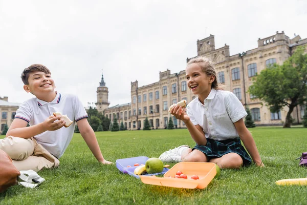 Niños multiétnicos positivos sosteniendo sándwiches cerca de loncheras en el césped - foto de stock