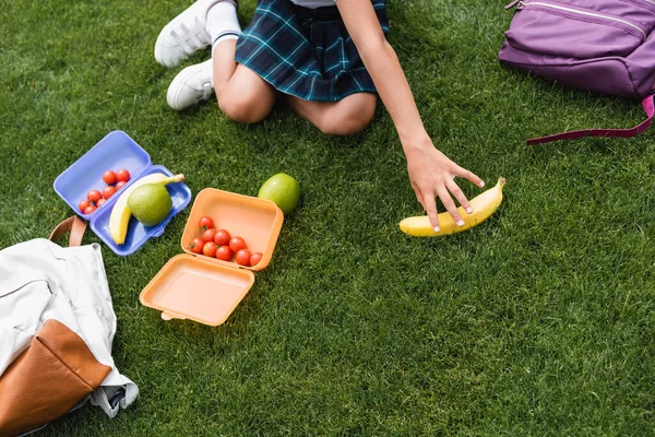 Cropped view of schoolgirl taking banana near backpacks and lunchboxes on grass — Stock Photo