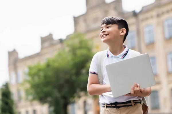Low angle view of smiling asian schoolboy holding laptop outdoors — Stock Photo