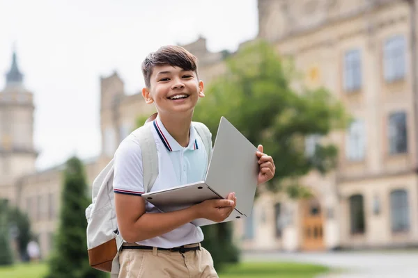 Cheerful asian schoolkid holding laptop and looking at camera outdoors — Stock Photo