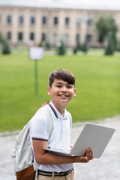 Happy preteen asian schoolboy with backpack holding laptop outdoors — Stock Photo