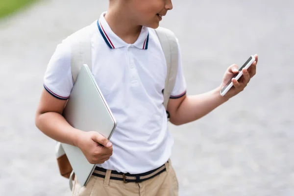 Cropped view of schoolboy holding smartphone and laptop outdoors — Stock Photo