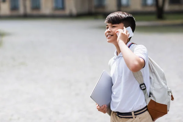 Lächelnder asiatischer Schüler hält Laptop in der Hand und telefoniert draußen — Stockfoto