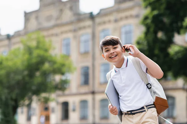 Веселый азиатский школьник держит в руках ноутбук и разговаривает по смартфону на улице — стоковое фото