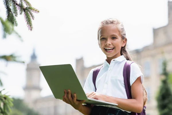 Feliz escolar sosteniendo el ordenador portátil y mirando a la cámara al aire libre - foto de stock