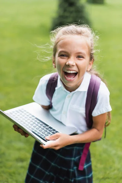 Cheerful schoolgirl looking at camera and holding laptop outdoors — Stock Photo