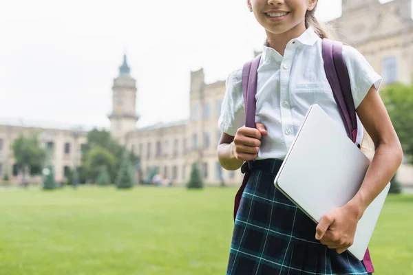 Vista recortada de la sonrisa preadolescente colegiala sosteniendo el ordenador portátil y la mochila al aire libre - foto de stock