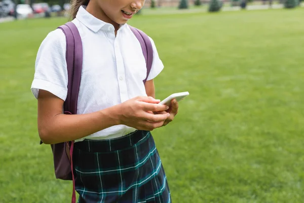 Обрезанный вид улыбающегося школьника с рюкзаком с помощью смартфона в парке — стоковое фото
