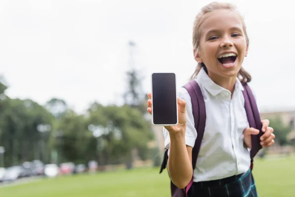 Весела школярка тримає смартфон з порожнім екраном в парку — стокове фото