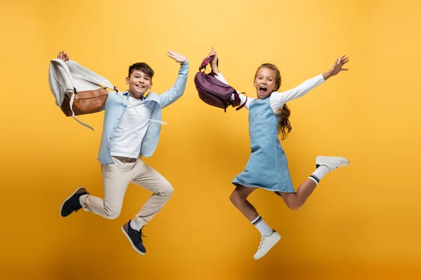 Aufgeregte multiethnische Kinder mit Rucksäcken springen auf gelbem Hintergrund — Stockfoto